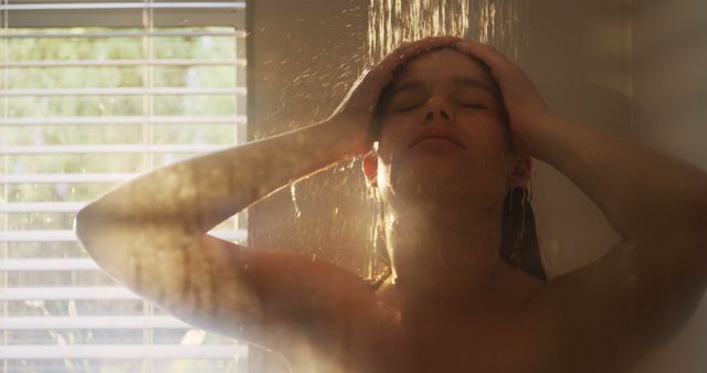 Young woman standing under shower with closed eyes and water cascading over her head. Sunlight is coming through window blinds, creating a serene and peaceful atmosphere. Perfect for concepts related to relaxation, hygiene, self-care, wellness, morning routines, and female lifestyle content.