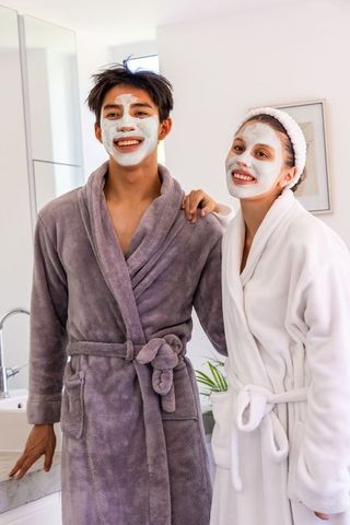 Smiling diverse couple with facial masks and plush bathrobes in bright bathroom enjoying pampering