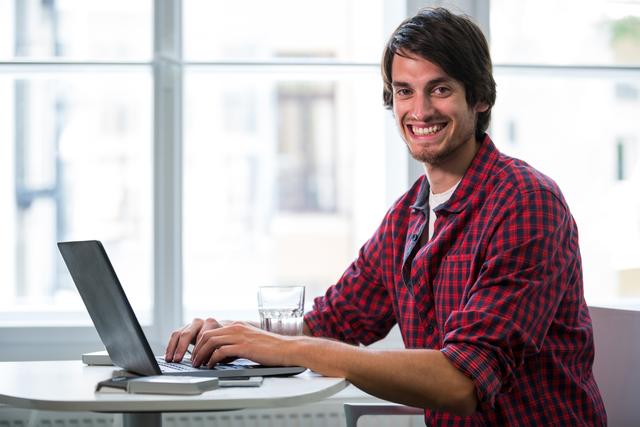 Young man in casual attire working on laptop in a modern office. Ideal for illustrating concepts of remote work, freelancing, modern business environments, productivity, and technology use in professional settings.