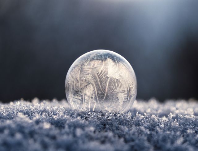 Close-up of a frosted soap bubble resting on snow on a sunny winter day. Ice crystals and patterns are visible within the bubble, creating a mesmerizing and intricate design. The scene captures the beauty of winter and frozen elements. Perfect for use in winter-themed campaigns, holiday cards, artistic prints, or nature blogs highlighting the intricacies of natural phenomena.