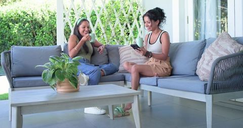 Friends sharing coffee and smartphone on covered patio sofa enjoying relaxed conversation