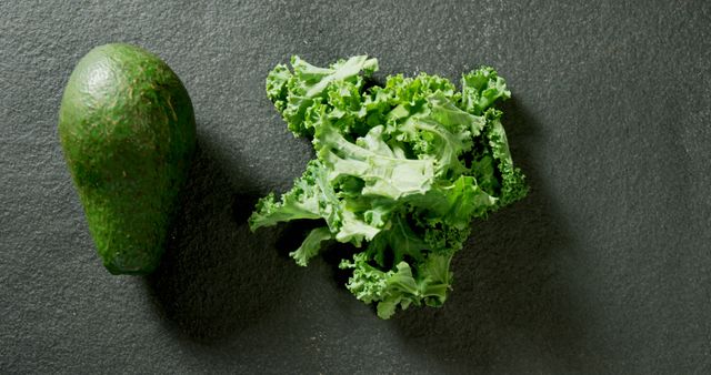 An avocado and a bunch of kale are placed on a dark slate surface, with copy space. These fresh ingredients are often used in healthy eating and vegetarian recipes.