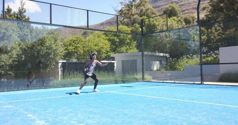 Fit woman playing padel on blue court swinging racket wearing visor and sunglasses