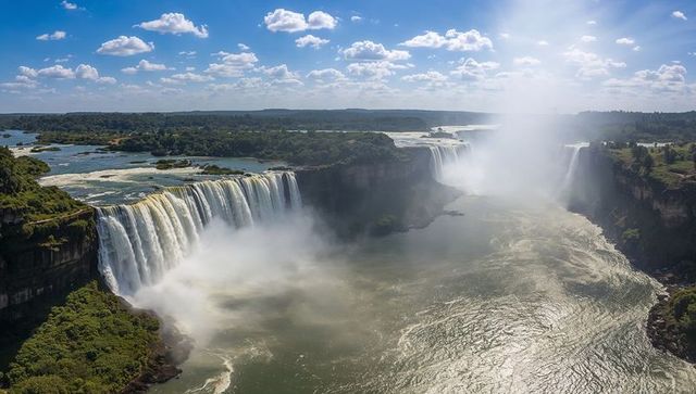 Majestic multi-segment waterfalls cascading into misty river gorge under sunlit blue sky