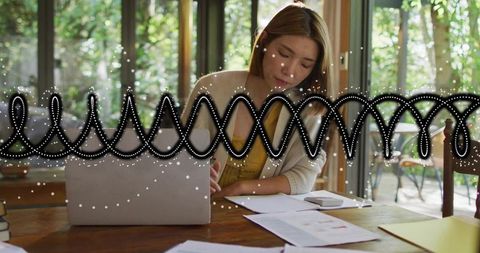 Remote worker writing notes at home desk with laptop, documents, sunlight and greenery