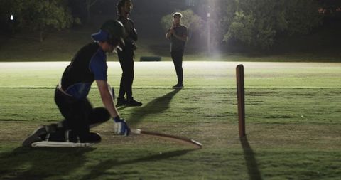 Kneeling batsman practicing near crease under floodlights with teammates watching at night