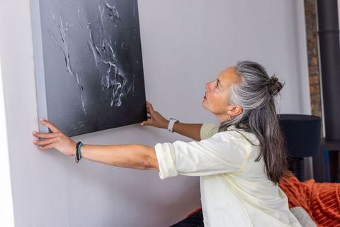 Senior woman adjusting dark abstract canvas on pale wall in cozy living room