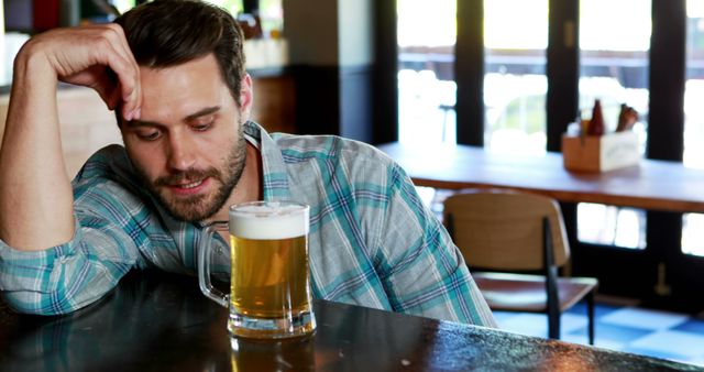 Man sitting at bar with a pint of beer, wearing a plaid shirt, looking pensive. Ideal for use in blogs or articles about relaxation, self-reflection, social drinking habits, or the ambiance of modern bars and pubs.