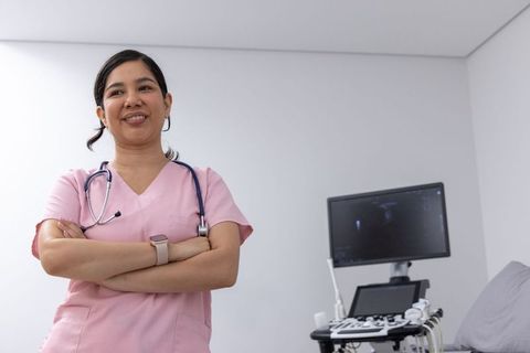 Confident clinician standing arms crossed in pink scrubs beside ultrasound unit