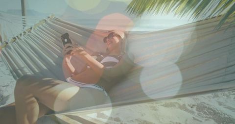 Sunlit woman relaxing in hammock on sandy beach, scrolling smartphone under palm shade