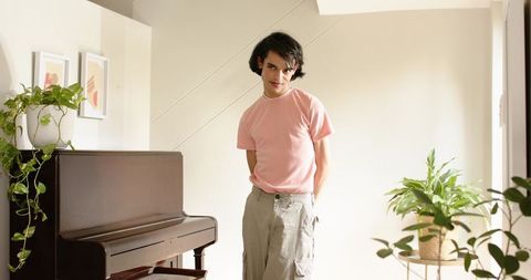 Young man standing beside upright piano in minimalist interior wearing pink tee in natural light