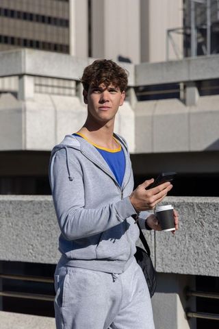 Young man in gray hoodie checking smartphone and holding coffee on urban rooftop