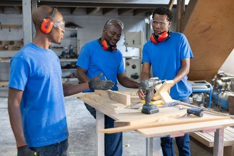 African american men collaborating on woodwork project in workshop wearing safety gear
