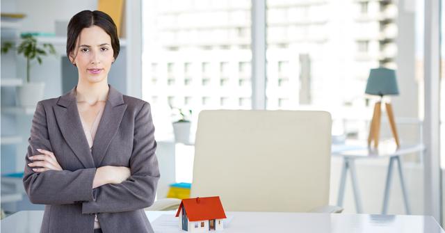 Female real estate agent standing confidently in modern office with arms crossed. Scale model of house on desk indicates realty business focus. Perfect for articles or promotions related to real estate, businesswomen, success in career, and the corporate world.