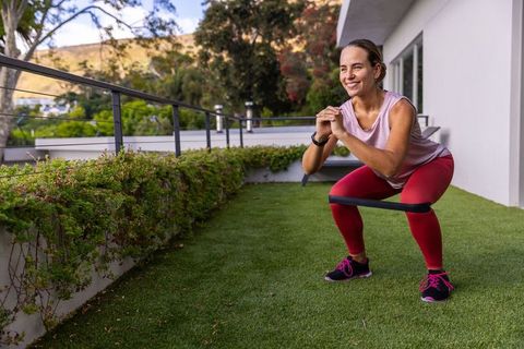 Smiling woman squatting with resistance band on rooftop terrace for home workout and strength traini