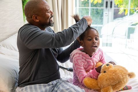 African american father brushing daughter hair on bed holding teddy bear morning routine