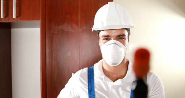 Confident construction worker wearing protective gear including a white hard hat and face mask. Ideal for use in articles, advertisements, or presentations related to construction safety, worker health, home renovation projects, and contractor services. Perfect for promoting occupational safety protocols and professional construction services.