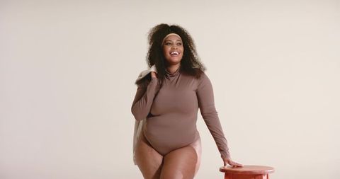 Confident african american woman posing in brown bodysuit with beige jacket and stool