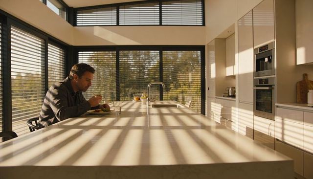 Man Eating at Sunlit Minimalist Kitchen Island with Long Marble Counter and Window Light