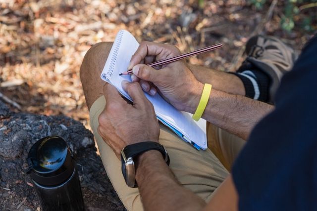 Man writing on notepad while resting on tree trunk in forest. Ideal for themes of outdoor activities, nature exploration, journaling, hiking, and relaxation. Useful for articles, blogs, and advertisements related to adventure, travel, and personal reflection.