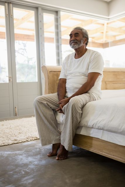 Senior man sitting on bed in a bright, modern bedroom, appearing thoughtful and relaxed. Ideal for use in articles or advertisements related to senior living, retirement, mental health, and peaceful home environments.