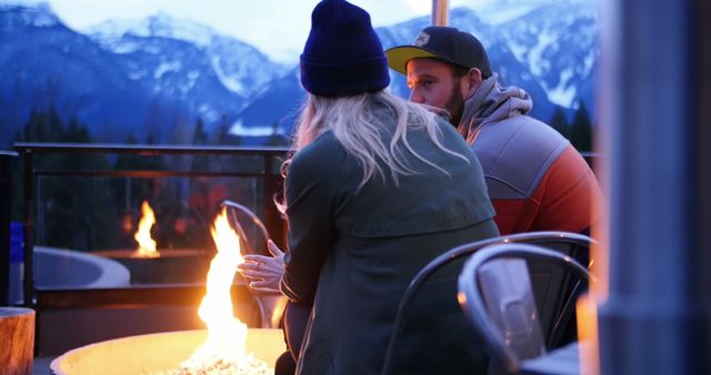 A Caucasian couple enjoys a romantic moment by a fire pit with a stunning mountain backdrop, with copy space. Their warm embrace and the cozy fire contrast with the chilly alpine setting, creating an intimate atmosphere.