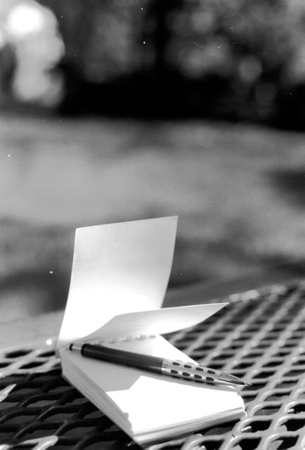 Open notebook with a pen resting on it, placed on a grid-like surface outdoors, captured in black and white. Suitable for themes related to writing, creativity, journaling, outdoor activities, and stationery. Perfect for blogs, articles, and advertising materials involving education, note-taking, and inspiration.