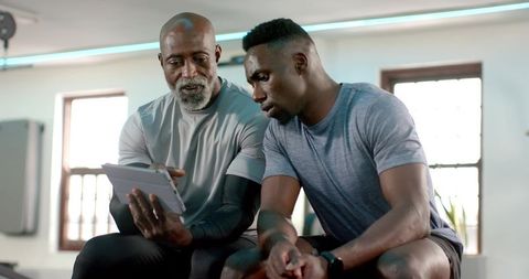 African american coach pointing at tablet with male trainee leaning on gym bench