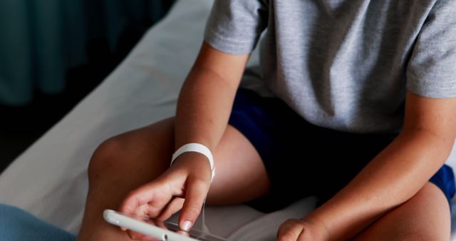 Child wearing hospital wristband, sitting on bed, and using smartphone. Depiction of a young patient using technology for entertainment or comfort during recovery. Useful for healthcare themes, pediatric care, or discussions about children's use of technology in hospitals.