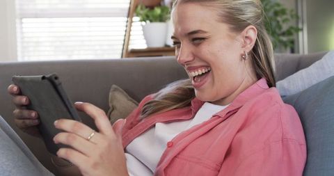 Laughing young woman relaxing on sofa using tablet for entertainment at home