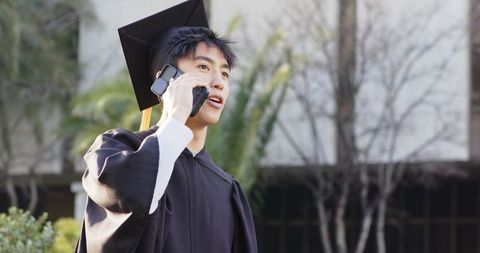 Young male graduate calling on smartphone in cap and gown on campus after commencement