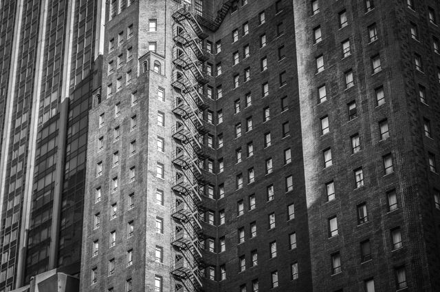 Dramatic black and white photo showing tall urban buildings with numerous windows and fire escapes. Perfect for use in articles on architecture, urban life, or city planning. Ideal for backgrounds, posters, and web designs featuring cityscapes or city life.