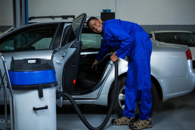 Mechanic in blue coveralls vacuuming car interior in a garage. Ideal for use in automotive service promotions, car maintenance advertisements, and professional cleaning service brochures.