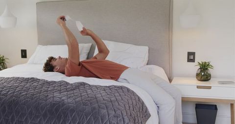 Relaxing young man lying on bed holding compact white device overhead in neutral bedroom