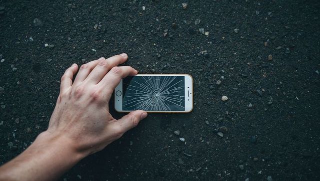Hand holding cracked smartphone on asphalt with scattered pebbles overhead