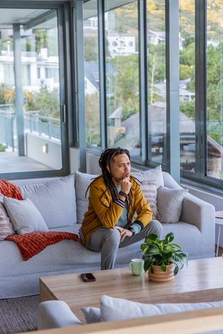 African american man sitting on gray sofa gazing out large windows in modern living room