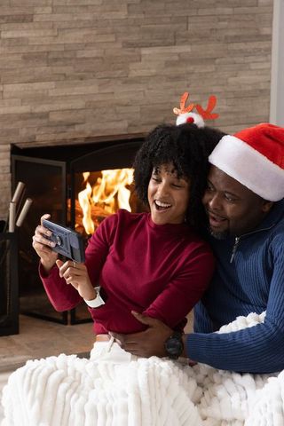 African american couple snuggling by fireplace taking festive selfie wearing santa hat