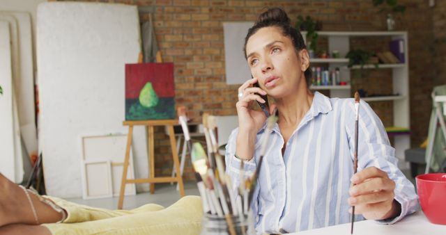 Female artist talking on phone while holding paintbrush in art studio. Scene features artist in striped shirt, sitting at desk with feet up, surrounded by paint brushes and art supplies. Brick wall background and casual atmosphere. Perfect for articles about creativity, artist's workspace, phone conversations, art-related professions, and inspirational work environments.