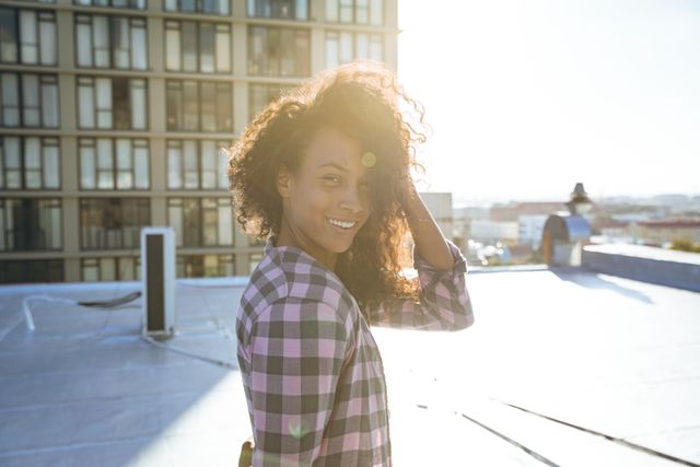 This image captures a happy biracial woman with long dark curly hair, wearing a checked shirt, smiling on an urban rooftop. The cityscape and sunlight create a modern and vibrant atmosphere. Ideal for use in lifestyle blogs, fashion websites, advertisements promoting urban living, or social media posts celebrating diversity and positivity.