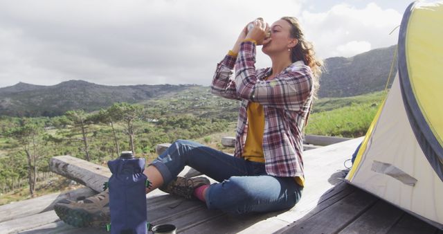 Woman camping in mountain area drinking coffee, sitting near tent. Ideal for use in travel blogs, outdoor gear promotions, adventure lifestyle content, advertisements, and marketing related to nature excursions and hiking.