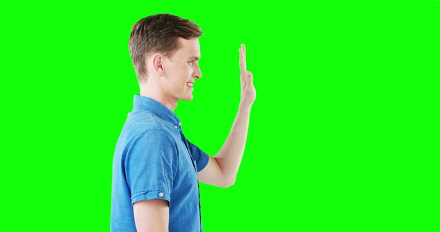 A man standing sideways in a blue shirt, smiling and waving with his hand against a green chroma key background. Ideal for use in video production, visual effects, and digital editing to insert diverse backdrops and create engaging content.
