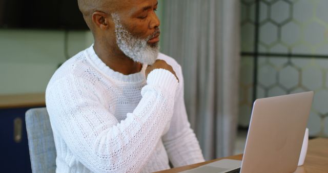 Senior man in white sweater looking uncomfortable with hand on shoulder, sitting at home office desk, indicating pain or injury. This can be used in articles about health, remote work ergonomics, or senior care. It is also useful for illustrating the impact of prolonged computer use among older adults.