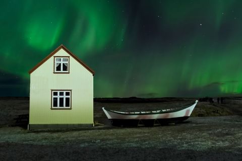 Northern Lights Dancing Over Coastal House and Beached Boat at Night in Rural Iceland