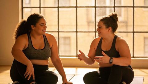 Women chatting on yoga mats in sunlit loft, smiling, relaxing, wearing smartwatch