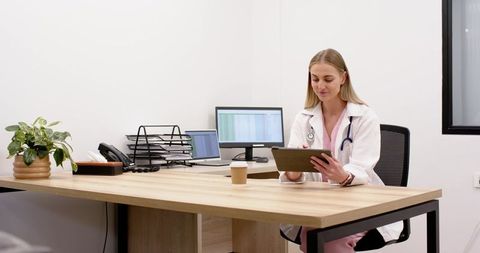 Female clinician reviewing tablet at clinic desk with monitor and laptop