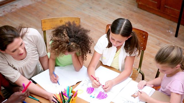 Teacher and students engaging in a collaborative drawing activity in an elementary school setting. Various art supplies like colored pencils are visible on the table. This image can be used for educational publications, school brochures, or children's creative programs emphasizing teamwork and creativity in learning environments.