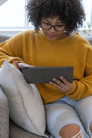African american woman sitting on gray sofa using tablet, wearing mustard sweater
