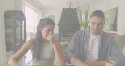 Couple Sitting at Table Looking Thoughtful in Sunlit Living Room with Fireplace and Plants