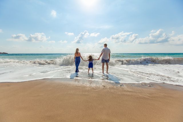 A family, consisting of parents and a child, is enjoying quality time together at the beach. They are holding hands while standing near the shoreline, with gentle waves crashing onto the sandy shore. The bright sun shines above, creating an idyllic and warm atmosphere. This image is perfect for illustrating family vacations, outdoor activities, leisure time, and the joy of spending time together in nature. It can be used for travel advertisements, tourism brochures, family-oriented marketing, and educational materials about family bonding.