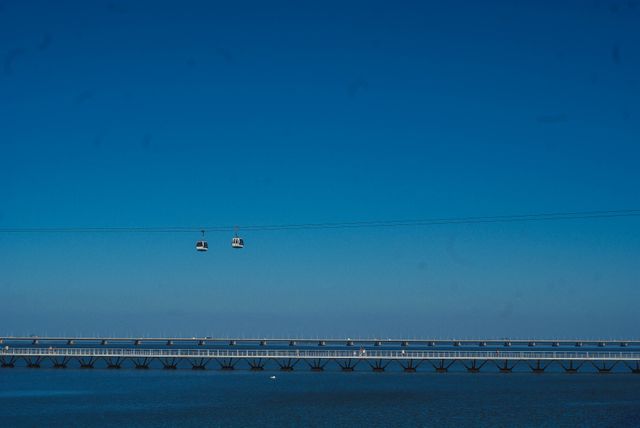 This image shows two cable cars moving across tranquil water against a clear blue sky backdrop with a bridge beneath them. The scene captures the serene and minimalistic beauty of transportation over water. Ideal for travel, transportation, infrastructure, and nature-related projects, this image promotes calmness and connectivity.